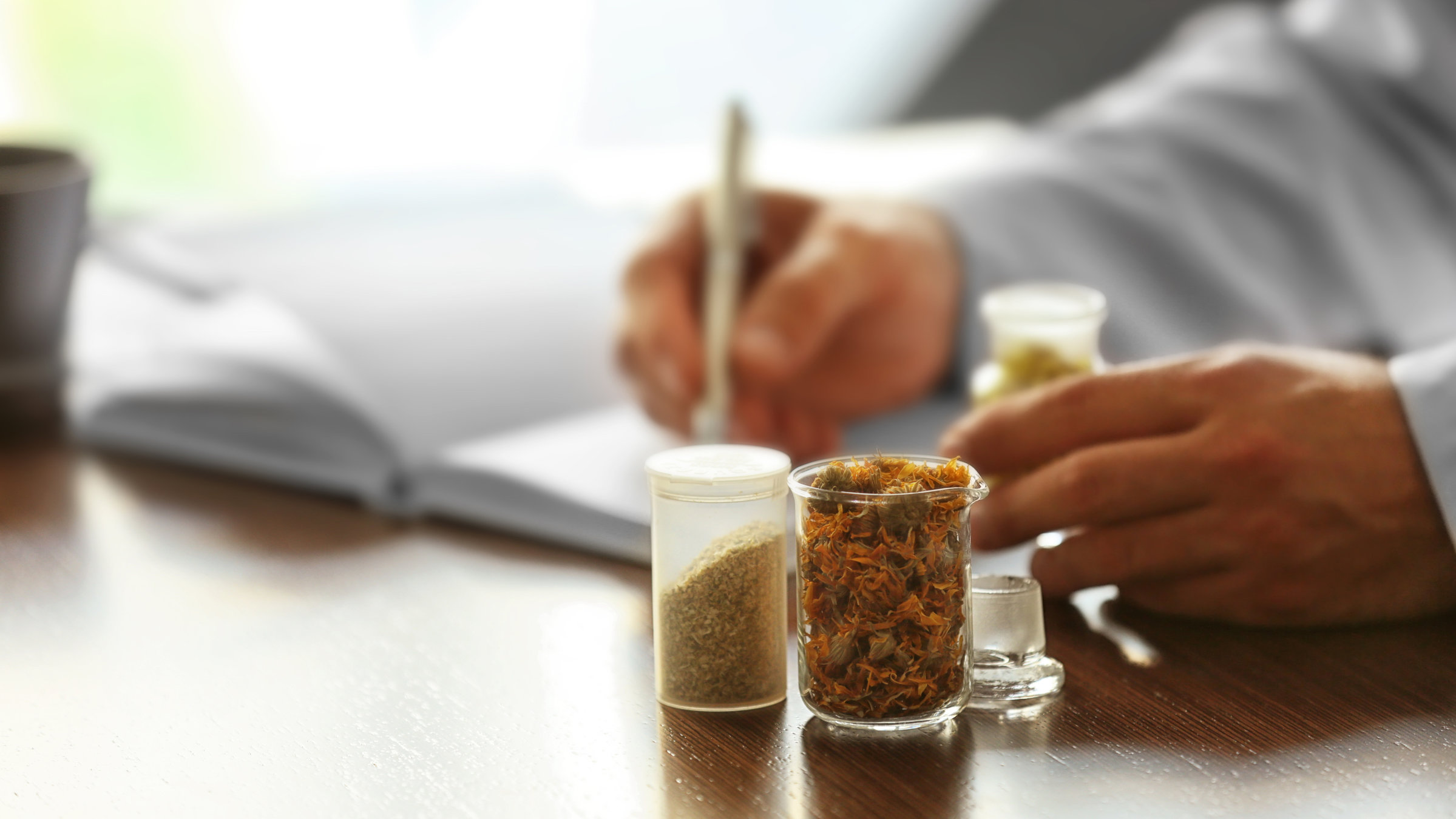 "37°: In den Fängen von Scharlatanen - Wenn aus Heilsversprechen Unheil wird": Auf einem Schreibtisch stehen zwei kleine Glasbehälter mit Kräutern. Im Hintergrund sitzt eine Mann, von dem nur die Hände zu sehen sind. Er schreibt etwas in ein Buch, während er einen Behälter mit gelbem Inhalt umfasst.