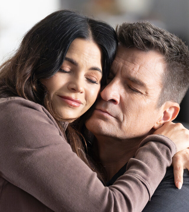 "The Rookie - Der letzte Tag": In einem Wohnbereich sitzt Officer John Nolan (Nathan Fillion) an einem Holztisch und Bailey (Jenna Dewan) auf seinem Schoß. Die beiden umarmen sich mit geschlossenen Augen.