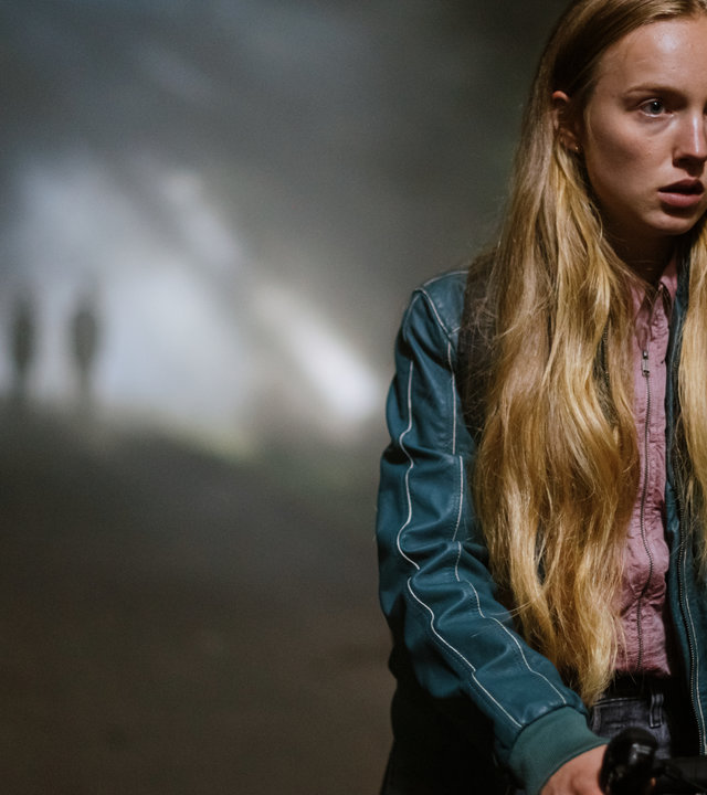 "Was wir fürchten - Familienbande": Lisa Abel (Mina-Giselle Rüffer) schiebt ihr Fahrrad durch den dunklen Wald. Sie schaut verängstigt nach vorne. Im Hintergrund stehen zweie Gestalten, die undeutlich zu erkennen sind.