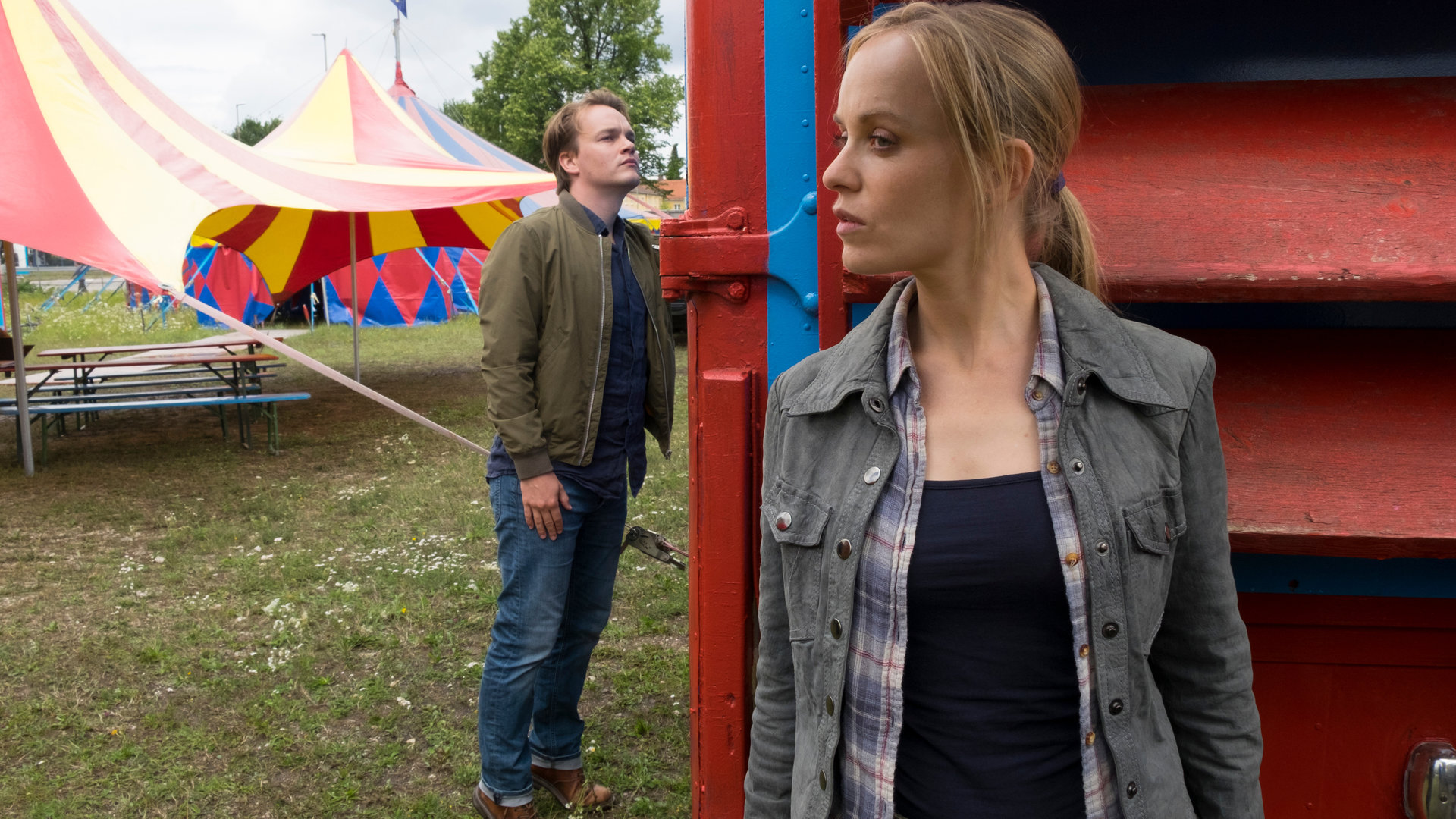 "Der Alte - Fürs Kind allein": Kathrin Weber (Friederike Kempter) versteckt sich hinter einem Wohnwagen, den Kommissar Tom Kupfer (Ludwig Blochberger) inspiziert. Im Hintergrund sind Zirkuszelte zu sehen.