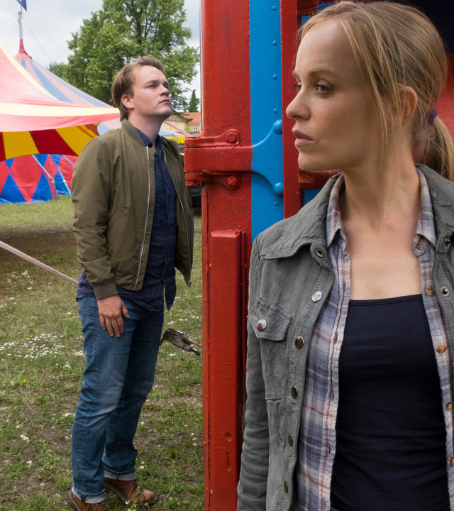 "Der Alte - Fürs Kind allein": Kathrin Weber (Friederike Kempter) versteckt sich hinter einem Wohnwagen, den Kommissar Tom Kupfer (Ludwig Blochberger) inspiziert. Im Hintergrund sind Zirkuszelte zu sehen.