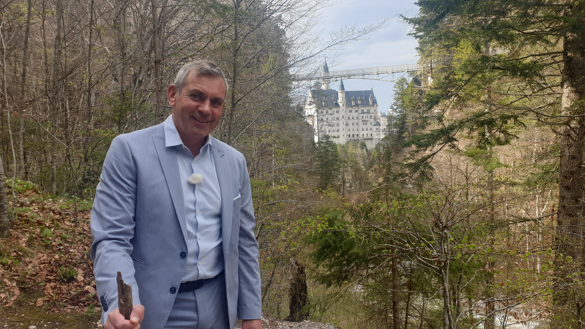 Kaminer steht im Wald, einen Wanderstock in der Hand. Im Hintergrund sieht man malerisch das Schloss liegen.