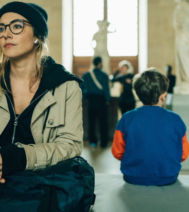 "Cat's Eyes - Prudence": Sylia (Constance Labbé) sitzt auf einer Bank in einem großen Raum im Louvre, in dem antike Statuen stehen. Mit dem Rücken zu ihr sitzt ein kleiner Junge neben ihr. Im Hintergrund, nur unscharf zu erkennen, befinden sich weitere Personen.