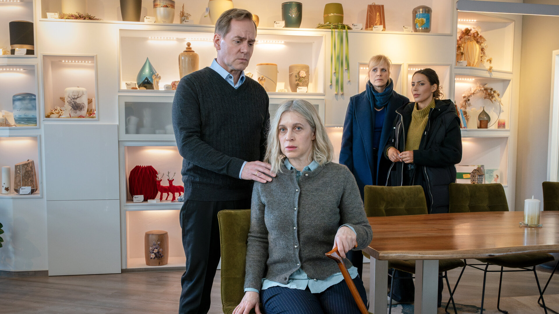 "SOKO Leipzig - Die letzten Tage": Michael Sonnenberg (Patrick Heyn) steht hinter seiner Frau Kathrin Sonnenberg (Nicole Ernst), die auf einem Stuhl am Kopf eines Tisches sitzt und sich zusätzlich auf einen Gehstock stützt. Michael hat seiner Frau eine Hand auf die Schulter gelegt. Ina (Melanie Marschke) und Kim (Amy Mußul) stehen im Hintergrund vor einer Wand auf der Urnen stehen und beobachten die beiden.