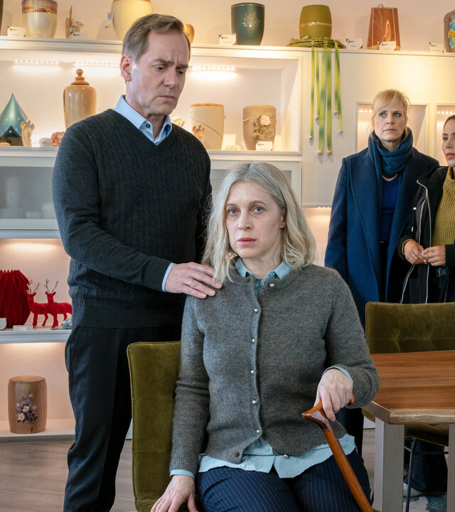 "SOKO Leipzig - Die letzten Tage": Michael Sonnenberg (Patrick Heyn) steht hinter seiner Frau Kathrin Sonnenberg (Nicole Ernst), die auf einem Stuhl am Kopf eines Tisches sitzt und sich zusätzlich auf einen Gehstock stützt. Michael hat seiner Frau eine Hand auf die Schulter gelegt. Ina (Melanie Marschke) und Kim (Amy Mußul) stehen im Hintergrund vor einer Wand auf der Urnen stehen und beobachten die beiden.