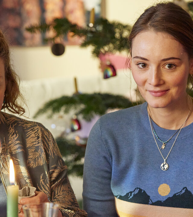 "Stille Nacht, raue Nacht": Svea (Victoria Fleer) und Liane (Maria Ehrich) sitzen vor einer festlich gedeckten Tafel nebeneinander und lächeln. Im Hintergrund steht ein Weihnachtsbaum.