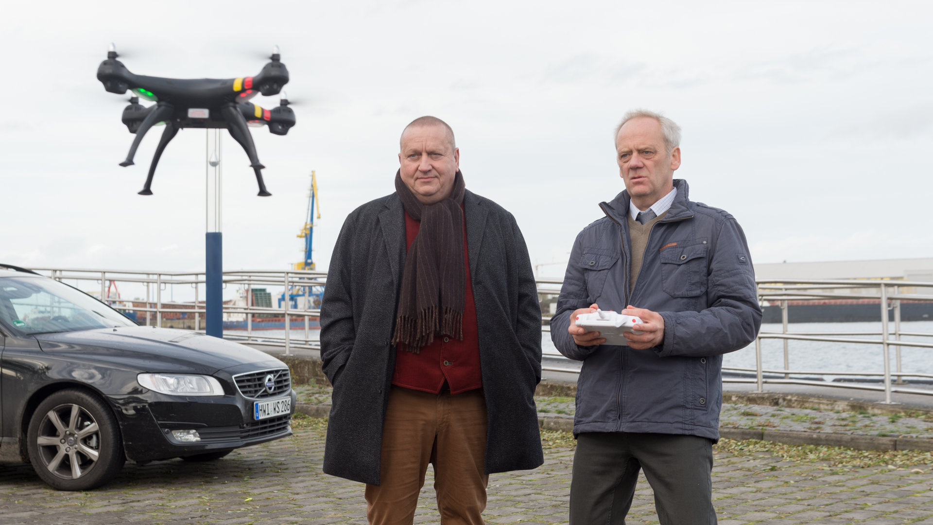 „SOKO Wismar – Geier Sturzflug“: Vor dem Wismarer Hafen steht Hauptkommissar Jan Reuter (Udo Kroschwald) mit KTU-Mann Johannes Hoff (Hans-Heinrich Hardt). Vor ihnen schwebt eine Drohne in der Luft, die Hoff mit einer Fernbedienung steuert.