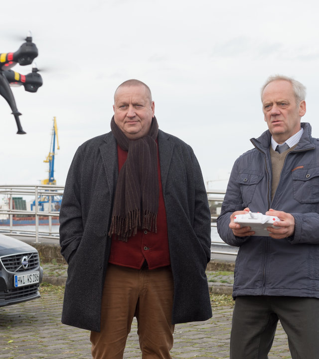 „SOKO Wismar – Geier Sturzflug“: Vor dem Wismarer Hafen steht Hauptkommissar Jan Reuter (Udo Kroschwald) mit KTU-Mann Johannes Hoff (Hans-Heinrich Hardt). Vor ihnen schwebt eine Drohne in der Luft, die Hoff mit einer Fernbedienung steuert.