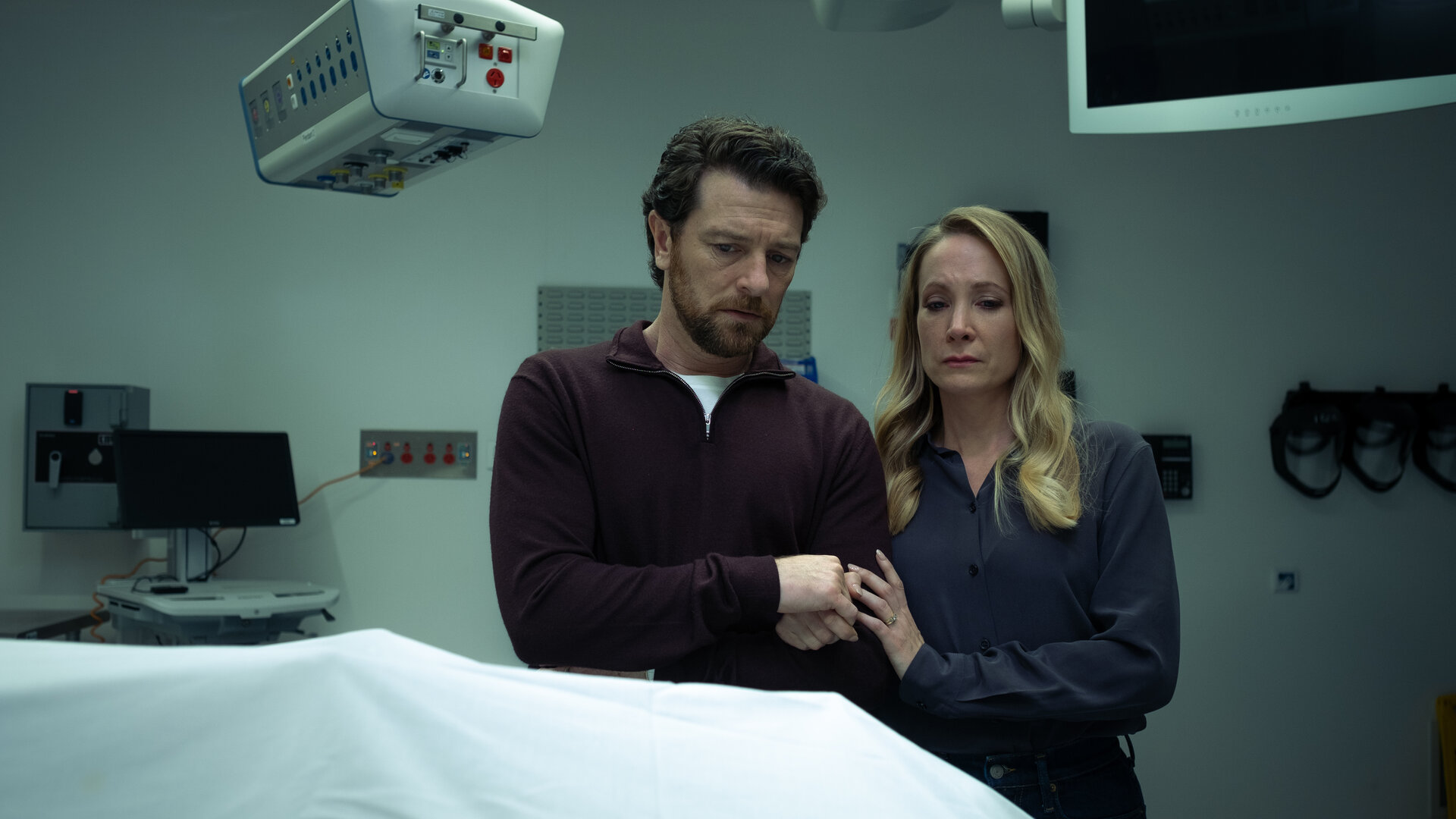 "North Shore – Tod in Sydney - Drummond Down Under": Abigail Crawford (Joanne Froggatt) und ihr Mann Simon Chalcott (Dan Spielman) stehen nebeneinander in einem Raum in der Gerichtsmedizin. Vor ihnen auf dem Tisch liegt eine Leiche unter einem weißen Tuch. Abigail hält Simons Hand.