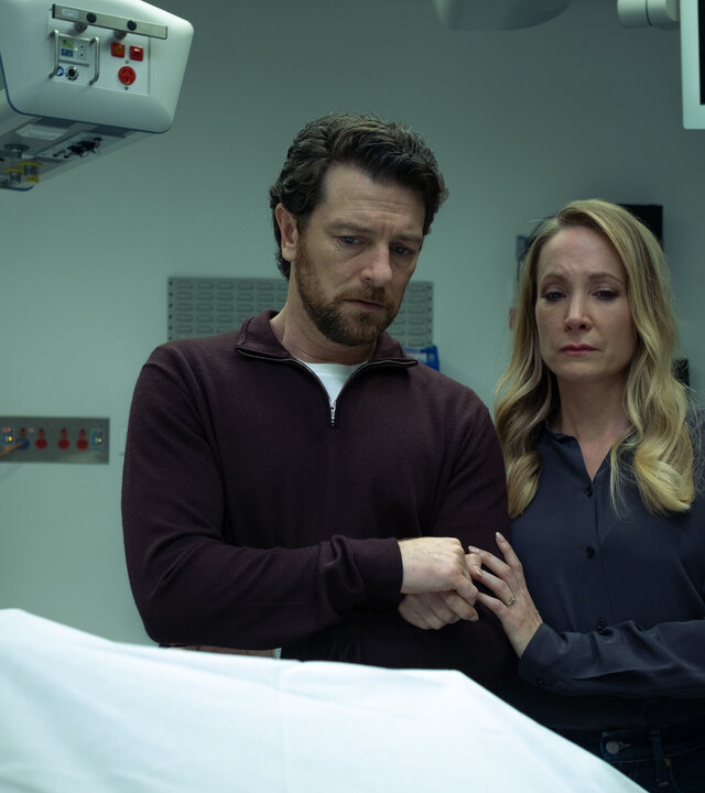 "North Shore – Tod in Sydney - Drummond Down Under": Abigail Crawford (Joanne Froggatt) und ihr Mann Simon Chalcott (Dan Spielman) stehen nebeneinander in einem Raum in der Gerichtsmedizin. Vor ihnen auf dem Tisch liegt eine Leiche unter einem weißen Tuch. Abigail hält Simons Hand.