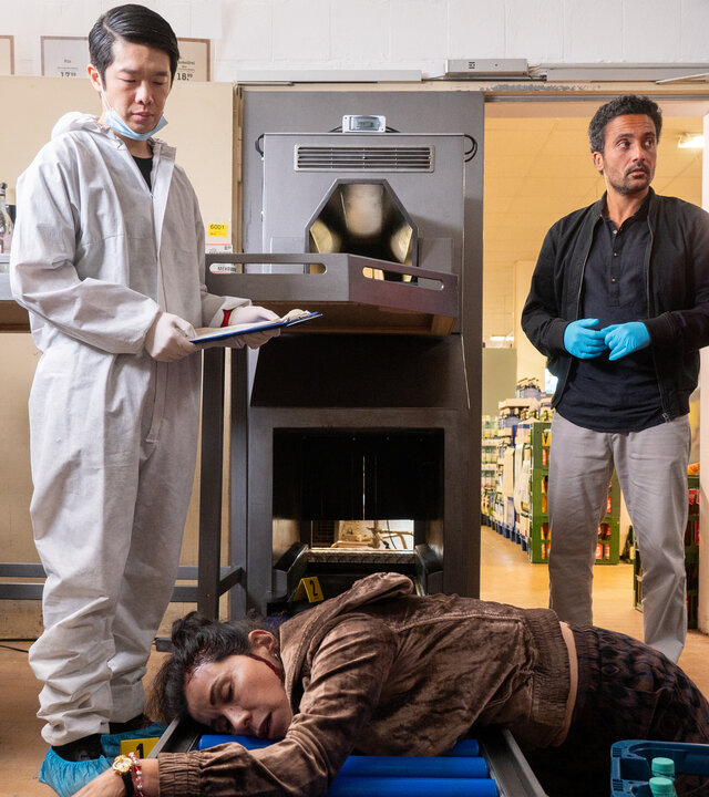 "SOKO Potsdam - Kassensturz": Pauline Hobrecht (Agnes Decker) und David Grünbaum (Omar El-Saeidi) stehen gemeinsam mit Spusi Thomas Brandner (Yung Ngo) am Tatort, im Lagerraum für Leergut des Supermarkts. Die Leiche der Frau (Komparsin) liegt auf einem Laufband für Leergut.