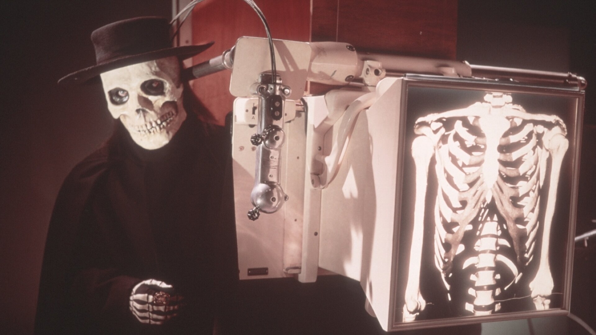 "Edgar Wallace: Im Banne des Unheimlichen": Ein Mann in einer Totenkopfmaske mit einem schwarzen Hut steht hinter einem medizinischen Gerät, das ein Röntgenbild seines Brustkorbs zeigt, auf dem ein Skelett deutlich zu erkennen ist.