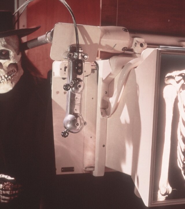 "Edgar Wallace: Im Banne des Unheimlichen": Ein Mann in einer Totenkopfmaske mit einem schwarzen Hut steht hinter einem medizinischen Gerät, das ein Röntgenbild seines Brustkorbs zeigt, auf dem ein Skelett deutlich zu erkennen ist.