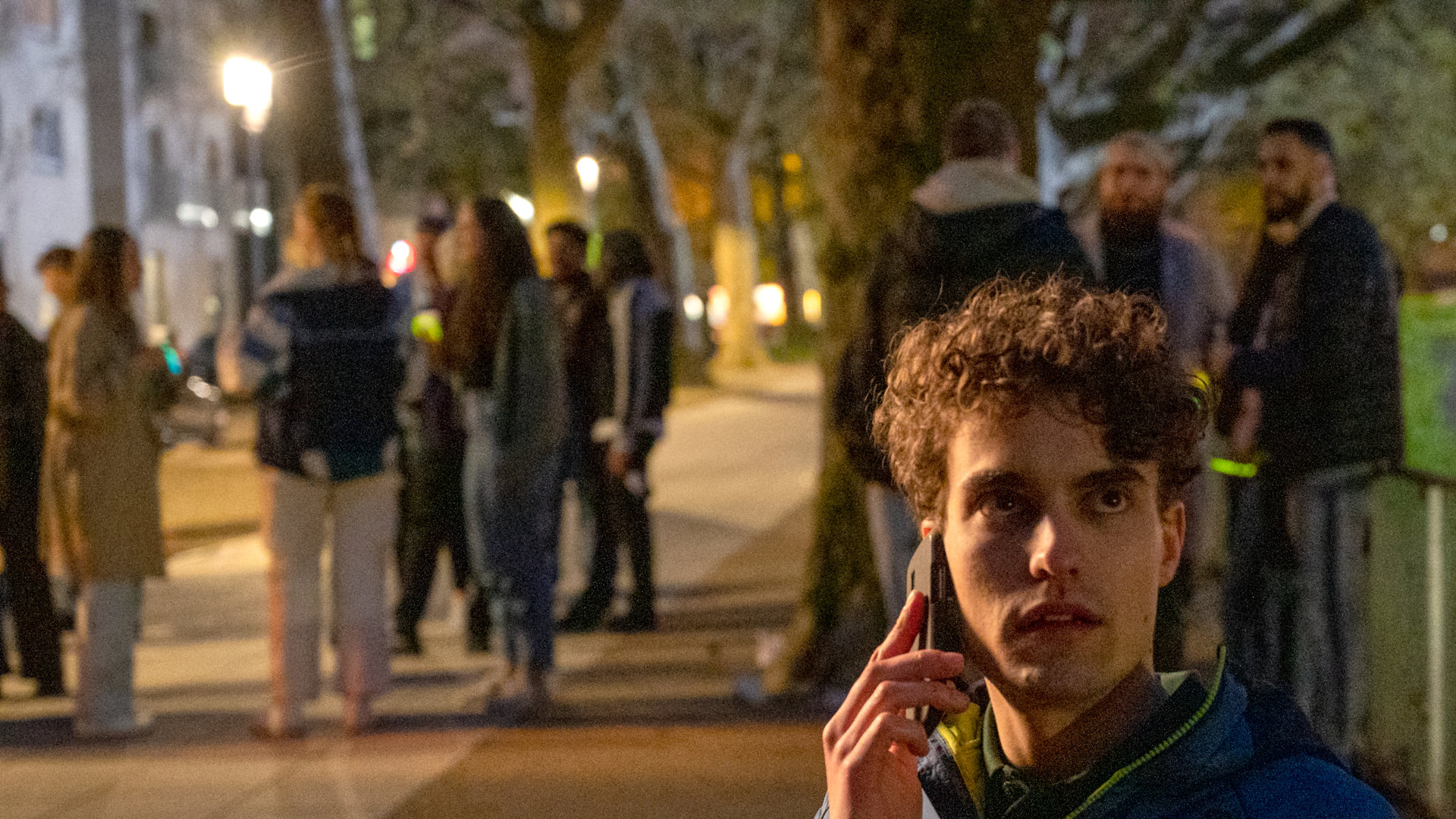 "SOKO Stuttgart - Partyzone": Marvin Kampfer (Lasse Möbus) sitzt bei Nacht auf einer Parkbank und hält sich mit der einen Hand sein Handy ans Ohr. In der anderen Hand hält er eine Bierflasche. Sein Blick ist erschrocken zur Seite gerichtet. Im Hintergrund stehen junge Menschen (Komparsen) mit Getränkeflaschen und unterhalten sich.