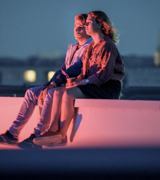 "SOKO Linz - Schöpfung 2.0": Niko Köhler (Felix Stichmann) und Lea Hoffmann (Sophie Bauer) sitzen bei Nacht auf dem Dach der Kepler-Universität. Indirekt werden sie von einem roten Licht angeleuchtet. Im Hintergrund sieht man einen Teil der Stadt Linz.