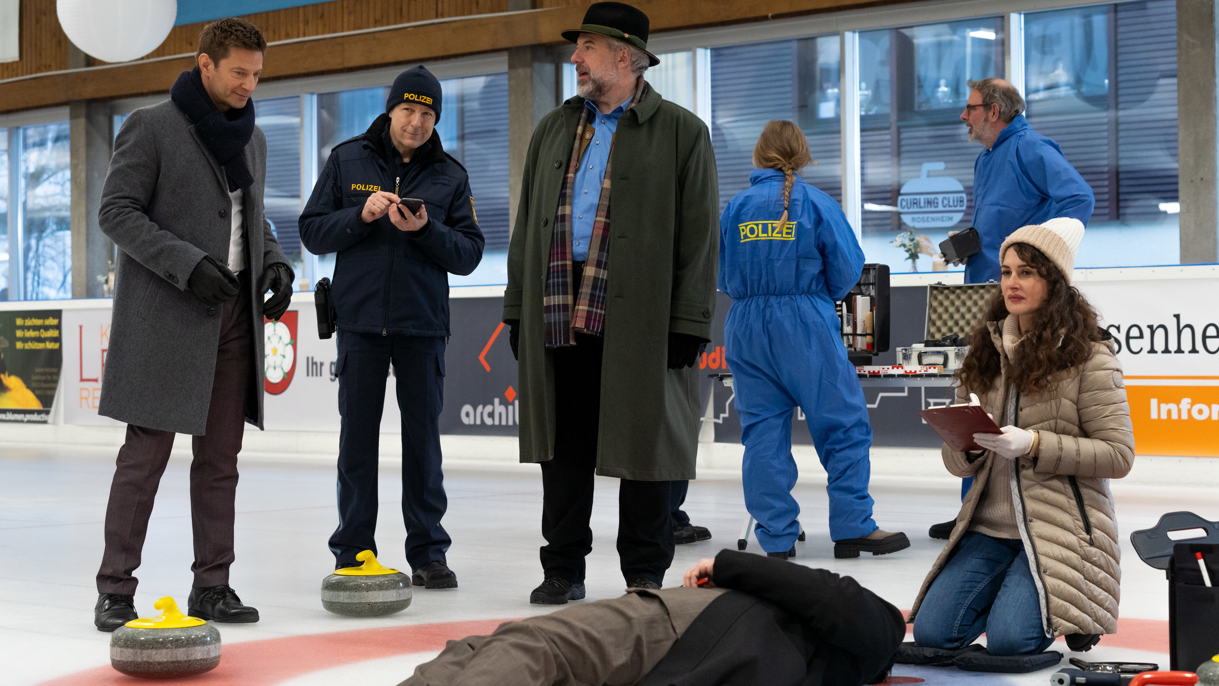 "Die Rosenheim-Cops - Ein eiskalter Mord": Kommissar Hansen (Igor Jeftić), Polizeihauptmeister Mohr (Max Müller), Kommissar Stadler (Dieter Fischer) sowie zwei Polizisten in blauer Schutzkleidung (Komparsen) stehen auf einer Eisfläche einer Halle. Um sie herum sind einige Curlingsteine mit gelben und roten Griffen zu sehen. Vor den Ermittlern liegt das Opfer auf dem Eis, daneben kniet Rechtsmedizinerin Dimos (Anastasia Papadopoulou), die eine Kladde in den Händen hält.