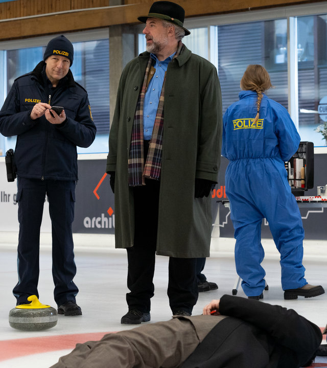 "Die Rosenheim-Cops - Ein eiskalter Mord": Kommissar Hansen (Igor Jeftić), Polizeihauptmeister Mohr (Max Müller), Kommissar Stadler (Dieter Fischer) sowie zwei Polizisten in blauer Schutzkleidung (Komparsen) stehen auf einer Eisfläche einer Halle. Um sie herum sind einige Curlingsteine mit gelben und roten Griffen zu sehen. Vor den Ermittlern liegt das Opfer auf dem Eis, daneben kniet Rechtsmedizinerin Dimos (Anastasia Papadopoulou), die eine Kladde in den Händen hält.