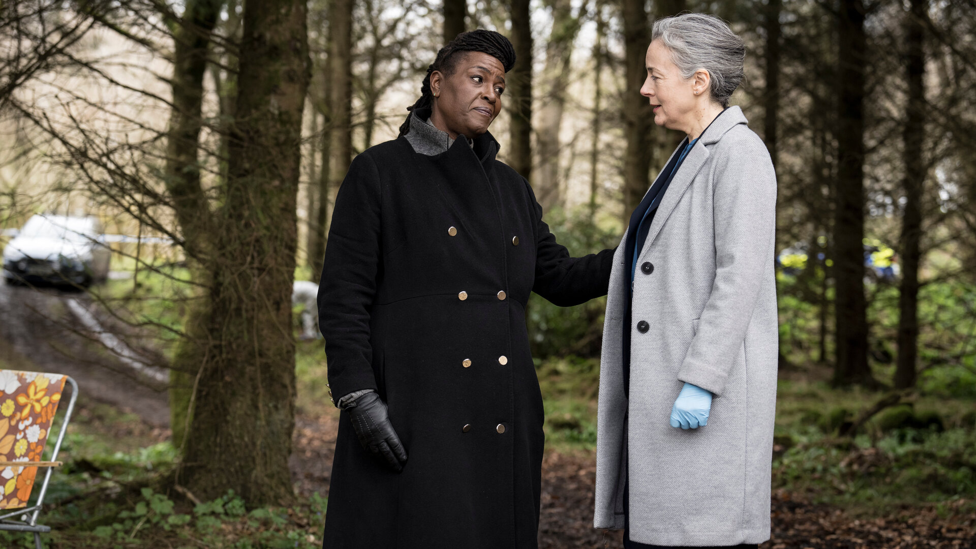 "Ellis - Am Abgrund": Ellis (Sharon D Clarke) und Val Cotton (Amanda Drew) stehen in einem Waldstück. Beide Frauen schauen sich an, Ellis hat ihre Hand beruhigend an den Arm von Cotton gelegt. Hinter ihnen parkt ein Auto auf einem Waldweg, an der Seite steht ein Campingstuhl.