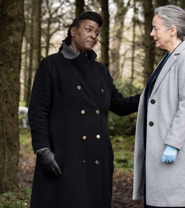 "Ellis - Am Abgrund": Ellis (Sharon D Clarke) und Val Cotton (Amanda Drew) stehen in einem Waldstück. Beide Frauen schauen sich an, Ellis hat ihre Hand beruhigend an den Arm von Cotton gelegt. Hinter ihnen parkt ein Auto auf einem Waldweg, an der Seite steht ein Campingstuhl.