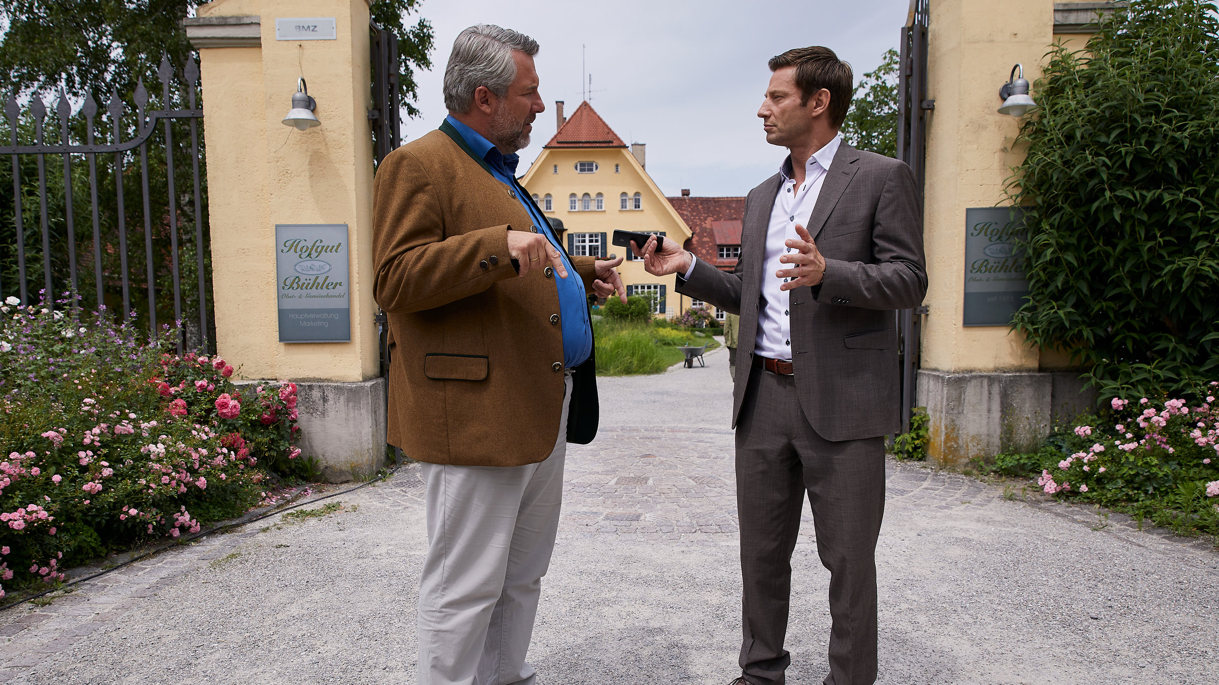 "Die Rosenheim-Cops - Drei Schwestern und ein Todesfall": Die Kommissare Stadler (Dieter Fischer) und Hansen (Igor Jeftic) stehen vor dem Eingangsportal des Hofguts Bühler und reden.