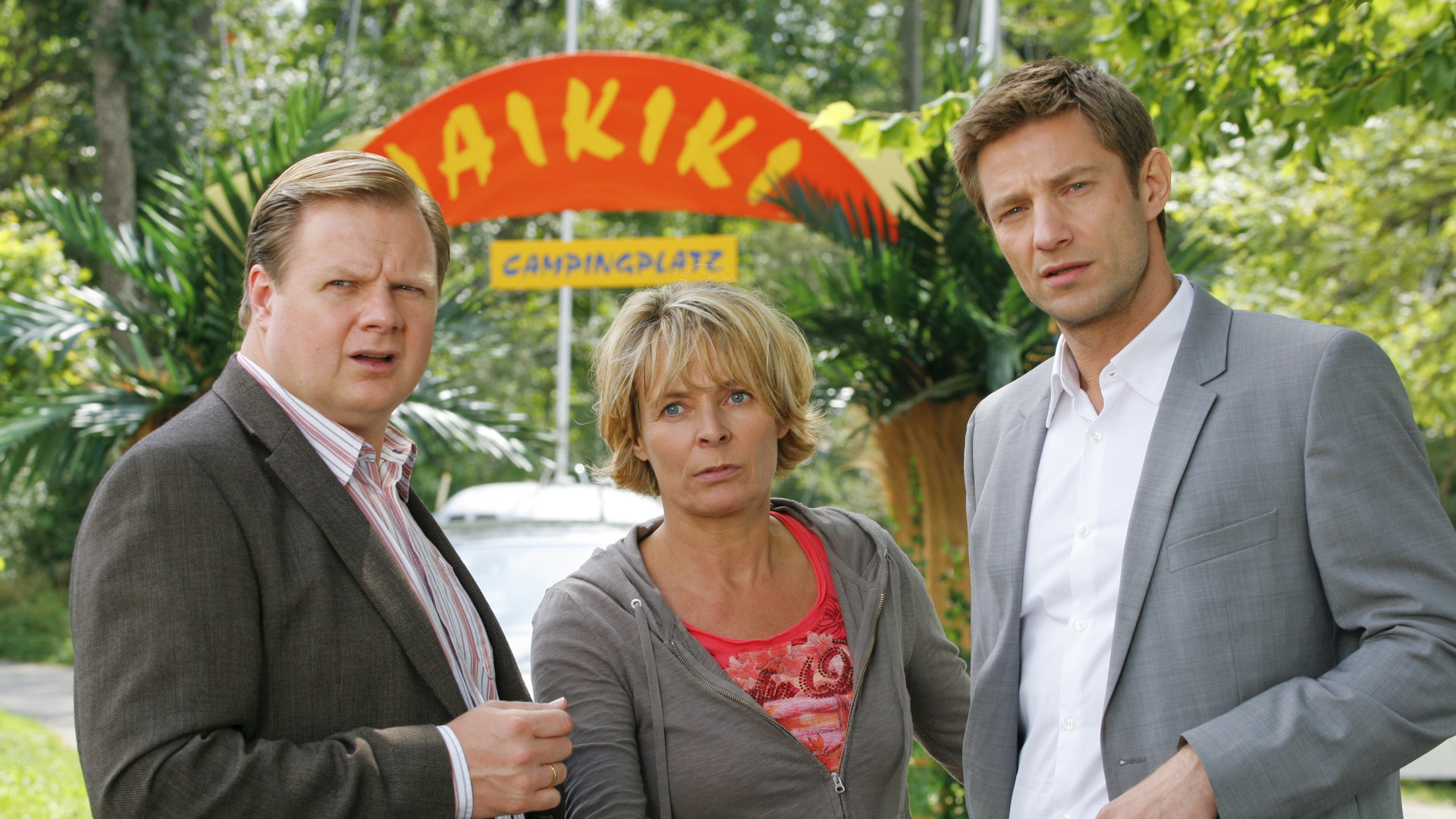"Die Rosenheim-Cops - Tod auf Waikiki": Auf dem Campingplatz "Waikiki" befragen die Kommissare Hansen (Igor Jeftic) und Hartl (Michael A. Grimm), Cornelia Meierling (Susanne Czepl). Im Hintergrund sind Bäume zu sehen sowie das Eingangsschild des Campingplatzes mit der Aufschrift "WAIKIKI CAMPINGPLATZ".