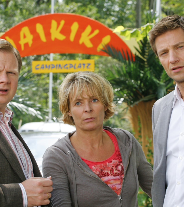 "Die Rosenheim-Cops - Tod auf Waikiki": Auf dem Campingplatz "Waikiki" befragen die Kommissare Hansen (Igor Jeftic) und Hartl (Michael A. Grimm), Cornelia Meierling (Susanne Czepl). Im Hintergrund sind Bäume zu sehen sowie das Eingangsschild des Campingplatzes mit der Aufschrift "WAIKIKI CAMPINGPLATZ".