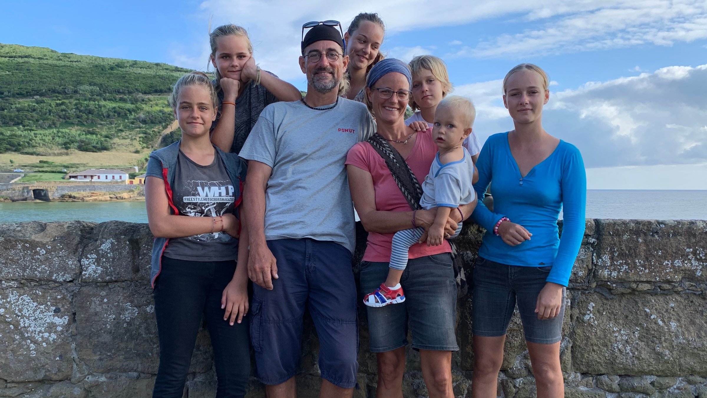 "37°: Aus und ahoi – Eine Großfamilie streicht die Segel": Die Großfamilie, Vater, Mutter und die sechs Kinder in von Kleinkind bis zum Jugendlichen stehen an einer Kaimauer. Im Hintergrund ist eine Meeresbucht und ein eine grüne Hügellandschaft zusehen.