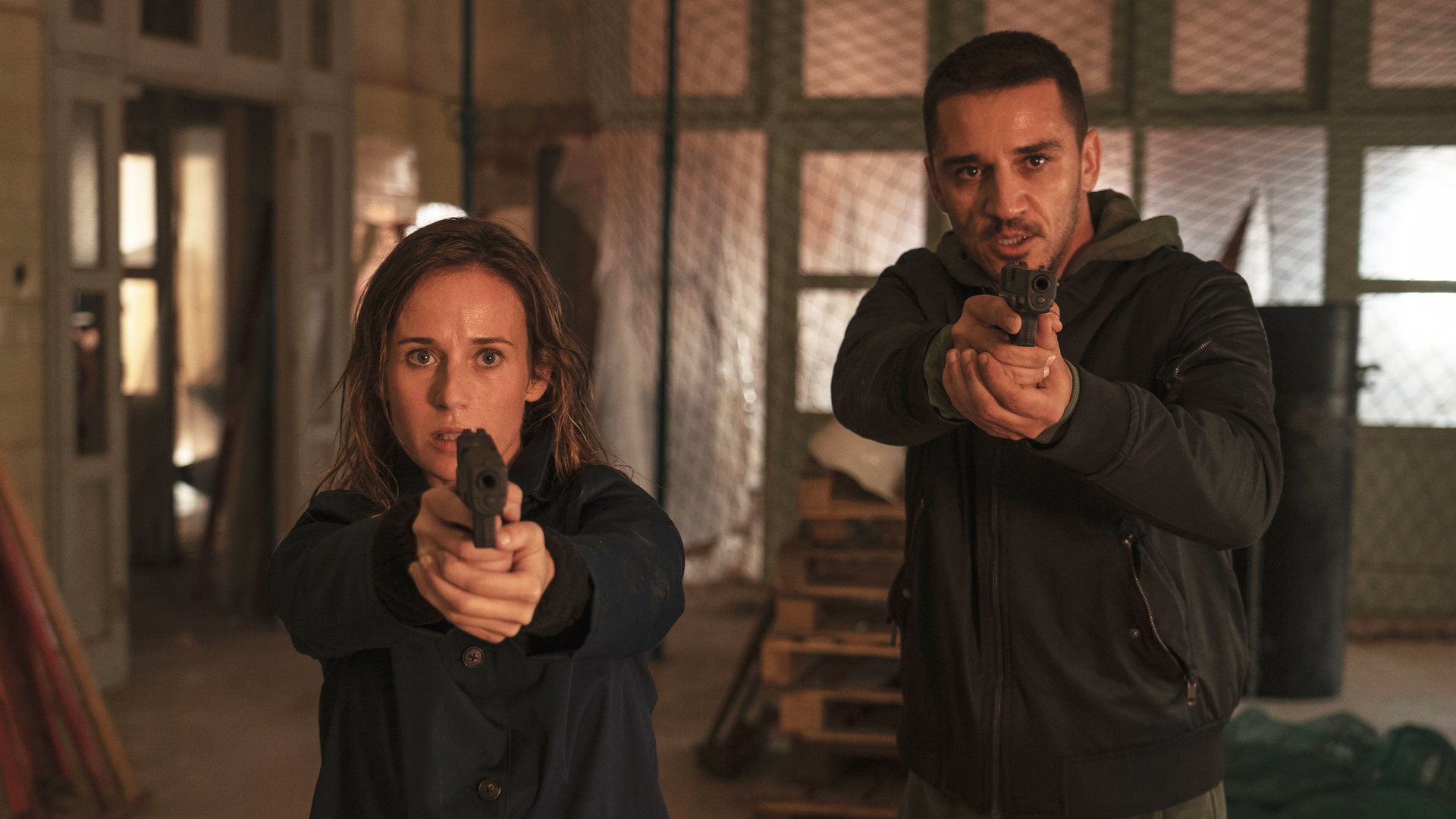 "Das Tal der vergessenen Kinder": Amaia Salazar (Marta Etura) und ihr Kollege Jonan Etxaide (Carlos Librado ''Nene'') stehen in einer Lagerhalle. Beide halten ihre Waffen vor sich und sehen angespannt in die Kamera.