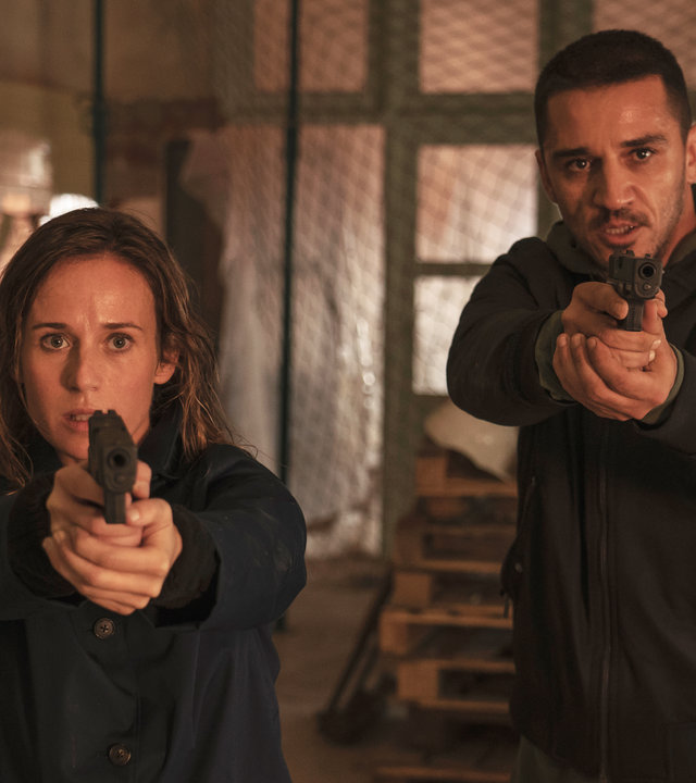 "Das Tal der vergessenen Kinder": Amaia Salazar (Marta Etura) und ihr Kollege Jonan Etxaide (Carlos Librado ''Nene'') stehen in einer Lagerhalle. Beide halten ihre Waffen vor sich und sehen angespannt in die Kamera.