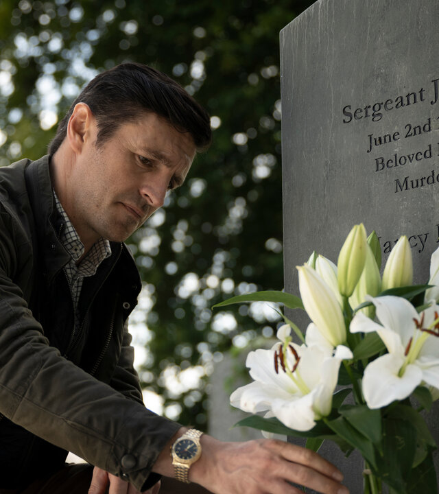 "Borderline – Grenzenlose Verbrechen - Das letzte Spiel": Detective Chief Inspector Philip Boyd (Eoin Macken) kniet vor einem Grabstein, auf dem die Namen seiner Eltern zu lesen sind. Er stellt gerade eine Vase mit weißen Lilien ab.