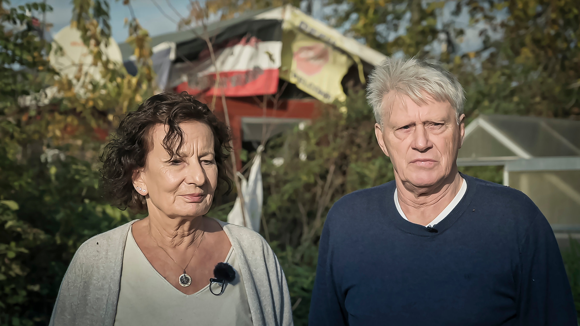"37°: Mein Nachbar, der Neonazi - Bleiben oder wegziehen?": Sylvia und Armin Joos stehen in ihrem Garten in Hörstgen (Nordrhein-Westfalen). Sylvia hat gelocktes kinnlanges Haar und trägt ein weißes T-Shirt, darüber eine beige Strickjacke. Armin hat graue Haare und trägt einen blauen Pulli. Im Hintergrund hängt ein Banner des Nachbarn mit den Aufschriften "Nationaler Widerstand" und "Refugees Not Welcome" sowie eine Schwarze Sonne. Diese Banner hängen an der Grenze zu Joos’ Garten und sind nur vom Ehepaar einsehbar.