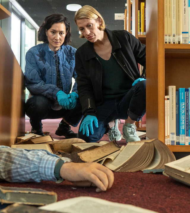 „SOKO Potsdam – Totgewälzt“: In einer hellen Hochschulbibliothek hocken Pauline Hobrecht (Agnes Decker) und Tamara Meurer (Anja Pahl) zwischen Bücherregalen am Boden. Vor ihnen liegt ein umgestürztes Regal, unter dem der Arm eines Mannes hervorragt. Zwischen verstreuten Büchern untersuchen die Kommissarinnen konzentriert den Tatort.