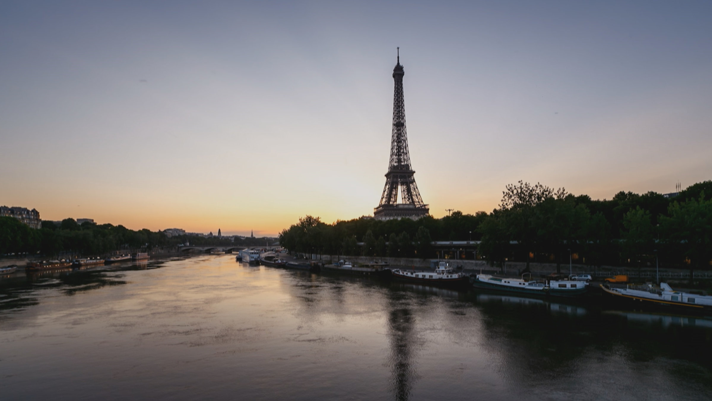 "Wunder der Baukunst (2/3) - Wahrzeichen": Kurz vor Sonnenaufgang über der Seine, an beiden Ufern sind Kähne vertäut, hinterm Kai viele Bäume. Auf der rechten Uferseite - zentral im Bild – der Eiffelturm.