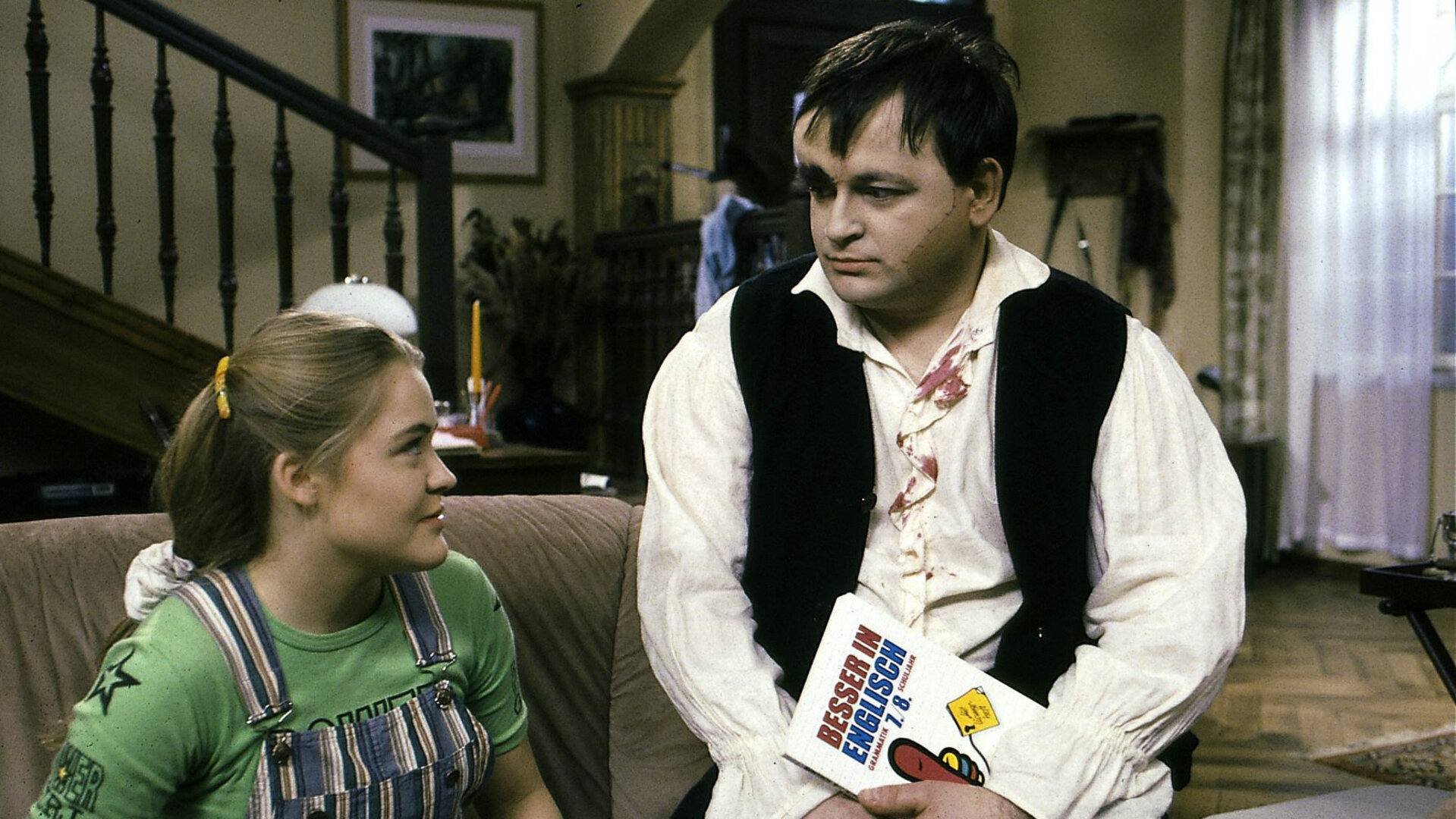 "Sprechstunde bei Dr. Frankenstein - English for Monsters": Franzi (Floriane Eichhorn) und Lenni (Michael Trischan) sitzen in einem Wohnraum auf dem Sofa und schauen sich an. Lenni hält ein Buch mit dem Titel "Besser in Englisch" in der Hand.