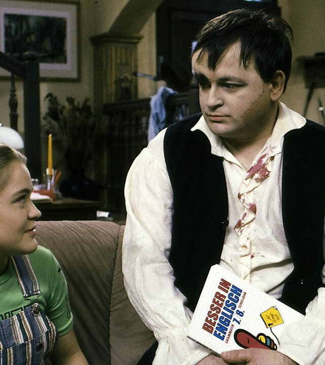 "Sprechstunde bei Dr. Frankenstein - English for Monsters": Franzi (Floriane Eichhorn) und Lenni (Michael Trischan) sitzen in einem Wohnraum auf dem Sofa und schauen sich an. Lenni hält ein Buch mit dem Titel "Besser in Englisch" in der Hand.