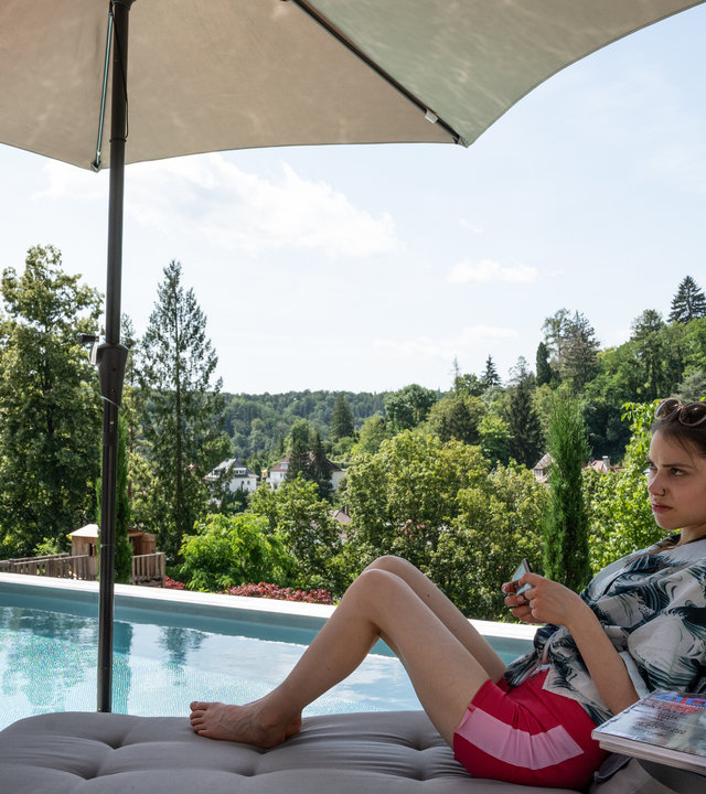 "SOKO Stuttgart – Sugar": Lara Hauser (Vivien Sczesny) sitzt in kurzer Hose und sommerlicher Bluse auf einer Sonnenliege. Über ihr ist einen Sonnenschirm aufgespannt, hinter ihr sieht man einen Pool und Bäume des Stuttgarter Talkessels. Neben der Liege steht ein Tischchen, auf dem ein Wasserglas, eine Zeitschrift, Kopfhörer und Lautsprecherboxen liegen. Kommissar Rico Sander (Benjamin Strecker) steht vor ihr und befragt sie zu der ermordeten Geliebten ihres Vaters. Lara schaut genervt.