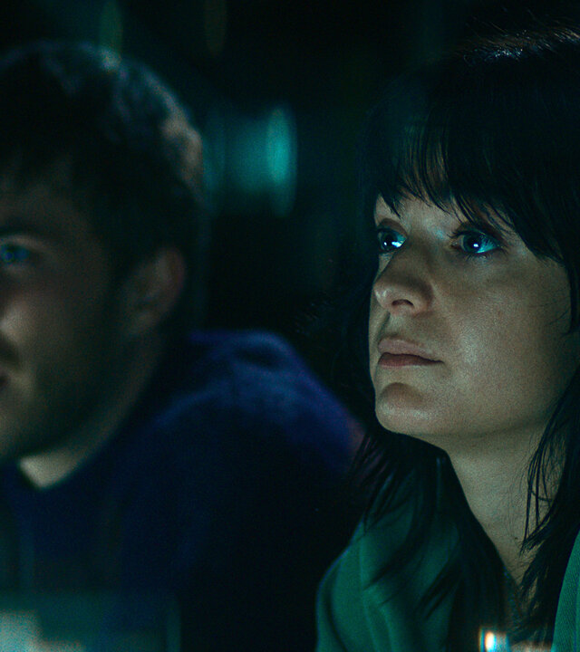 "The Kollective - Falsche Freunde": Aaron (Felix Mayr) und Delia (Céline Buckens) sitzen nebeneinander in einem dunklen Raum und beobachten etwas.