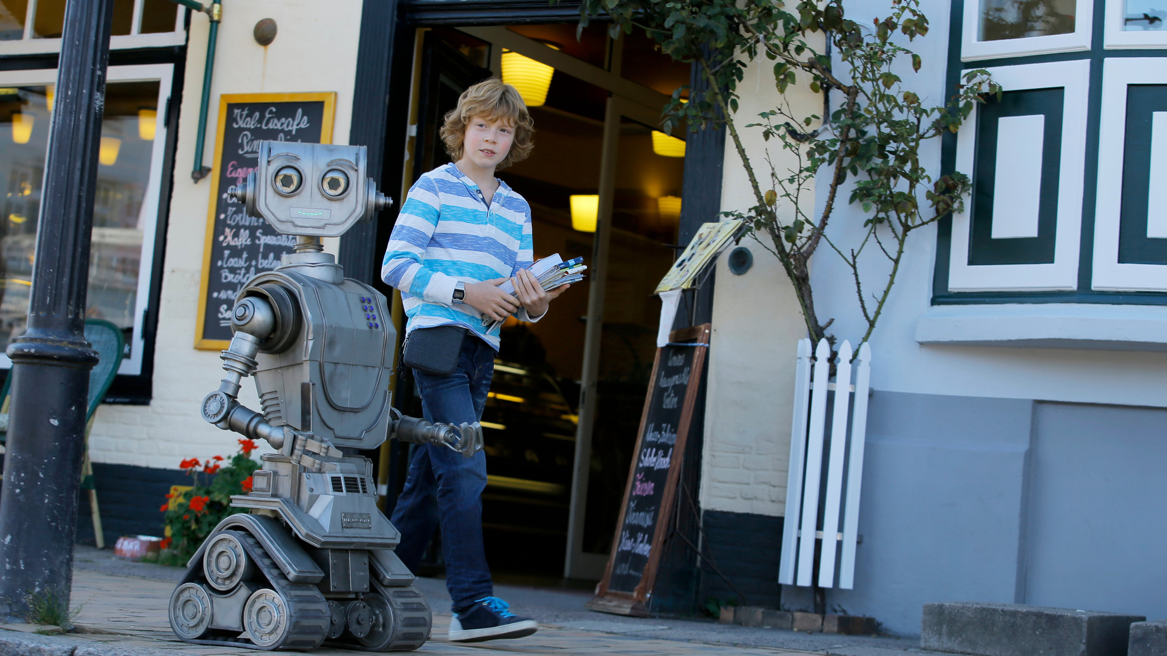 "Robbi, Tobbi und das Fliewatüüt": Tobbi (Arsseni Bultmann) und Roboter Robbi laufen beziehungsweise rollen nebeneinander die Straße entlang.