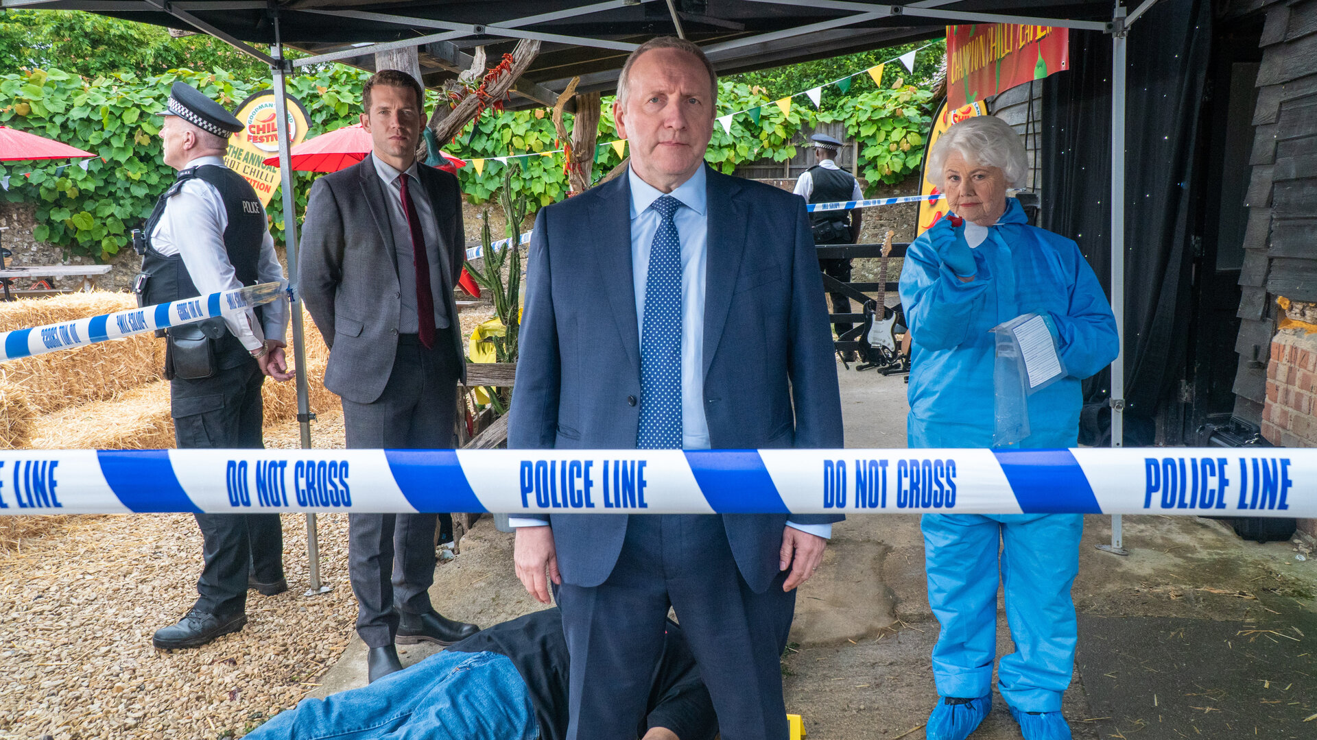 "Inspector Barnaby - Tödliches Klima": Sergeant Jamie Winter (Nick Hendrix), Inspector John Barnaby (Neil Dudgeon) und Rechtsmedizinerin Fleur Perkins (Annette Badland) stehen unter dem Vordach einer Hütte und schauen direkt die Kamera. Im Hintergrund liegt eine Leiche. Zwei Polizisten in Uniform (unbekannte Darsteller) bewachen den Fundort.