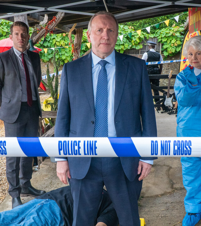 "Inspector Barnaby - Tödliches Klima": Sergeant Jamie Winter (Nick Hendrix), Inspector John Barnaby (Neil Dudgeon) und Rechtsmedizinerin Fleur Perkins (Annette Badland) stehen unter dem Vordach einer Hütte und schauen direkt die Kamera. Im Hintergrund liegt eine Leiche. Zwei Polizisten in Uniform (unbekannte Darsteller) bewachen den Fundort.