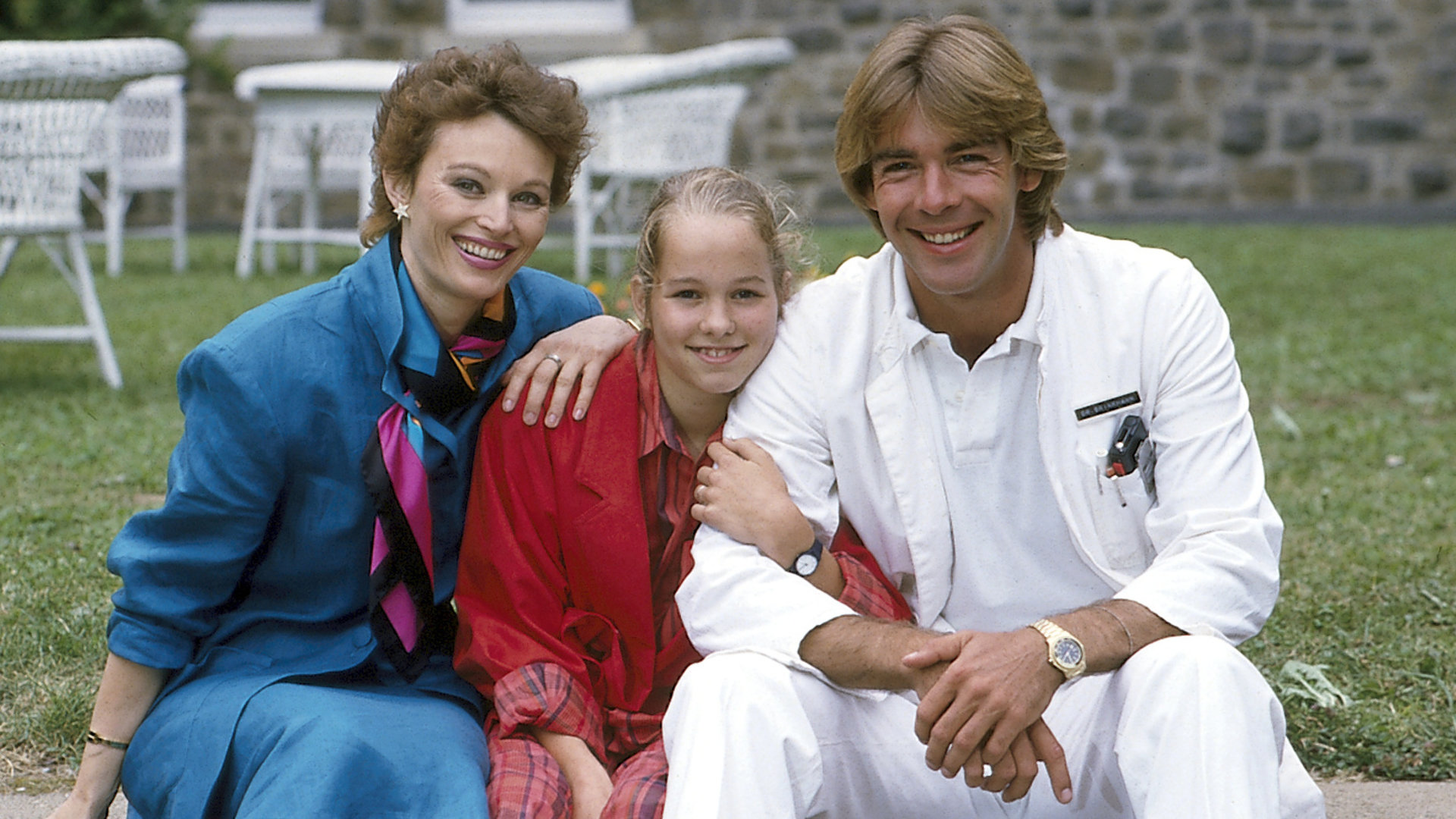 "Die Schwarzwaldklinik - Das Geständnis": Dr. Katarina Gessner (Ilona Grübel) Angie (Angelika Reißner) und Dr. Udo Brinkmann (Sascha Hehn) sitzen nebeneinander auf einer Mauer. Sie lächeln.