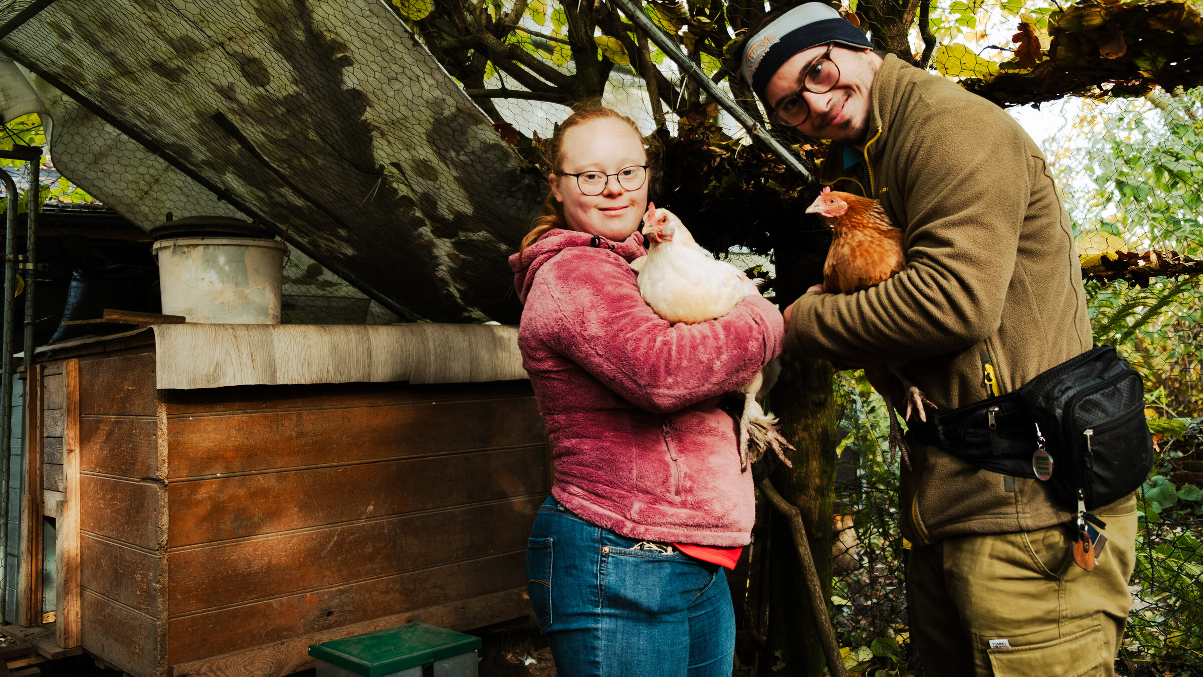 "Einfach Mensch - Teilhabe: Für alle schon Realität?": Hanna und Silas Gaub stehen im Garten, schauen lächelnd in die Kamera und halten beide ein Huhn im Arm.
