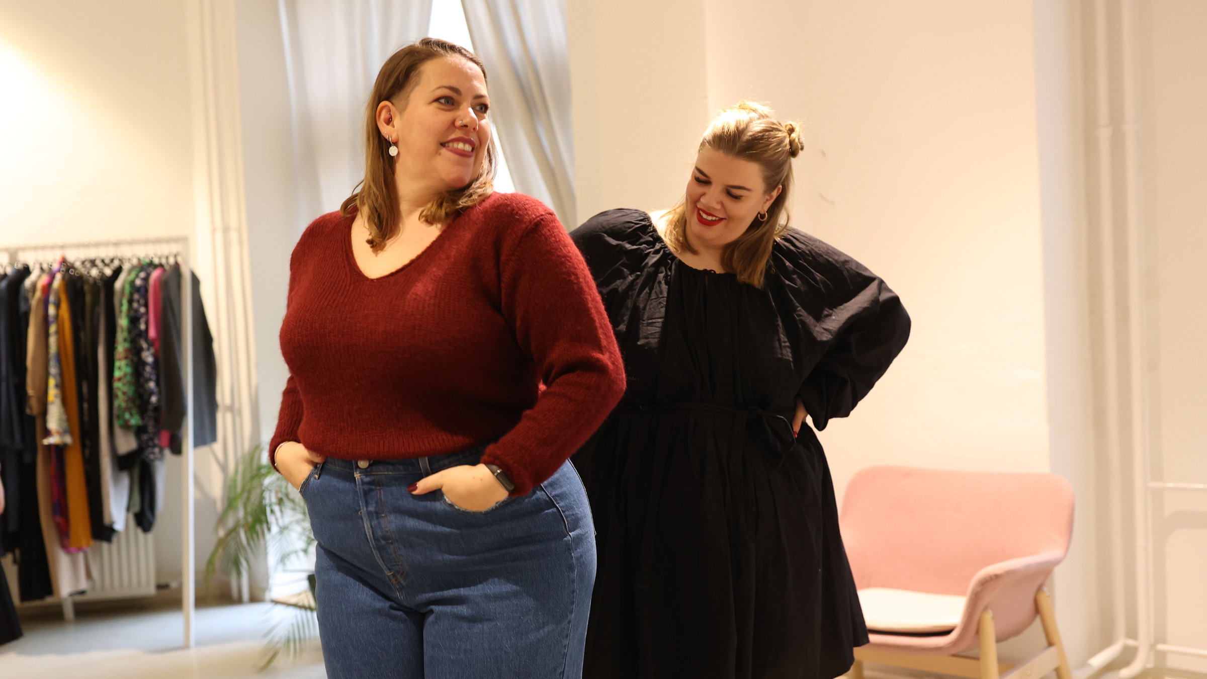 "37°: Glück kennt kein Gewicht - Mehrgewichtige und ihr neues Selbstvertrauen": Nicole, 39 Jahre alt, steht in der Bildmitte. Sie trägt Jens und einen roten Pullover, ihre dunkelblonden Haare sind halblang und am Oberkopf nach hinten gekämmt. Sie hat runde Ohrringe, ihre Lippen sind rot geschminkt. Hinter ihr steht eine junge blonde Frau, Mitte 20. Diese trägt ein elegantes schwarzes Mini-Kleid mit Puffärmeln. Sie guckt auf die andere Frau, die offensichtlich die Kleidungsstücke probiert und sich im Spiegel betrachtet, der jedoch außerhalb des Bildes ist.