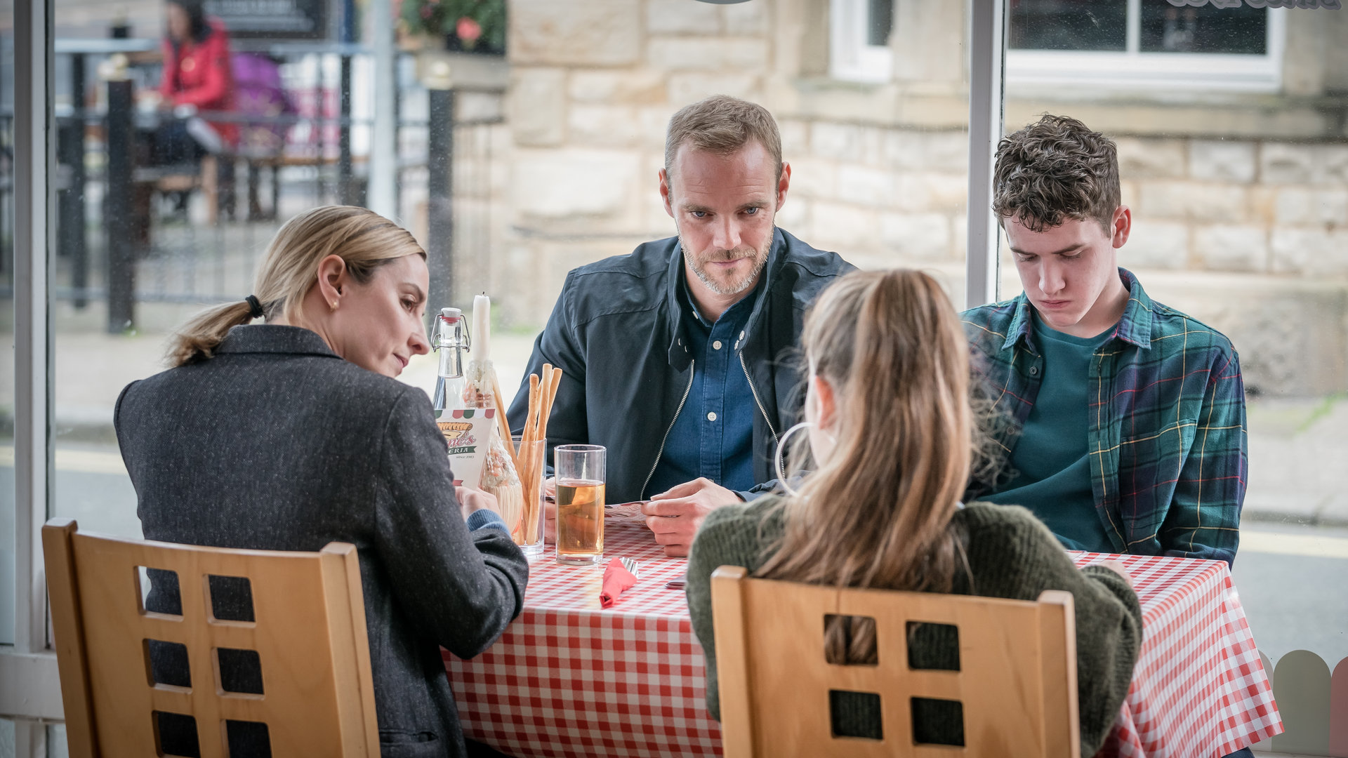 "The Bay - Vermutung": Lisa Armstrong (Morven Christie), Andy Warren (Joe Absolom), Rob Armstrong (Art Parkinson) und Abbie Armstrong (Imogen King) sitzen gemeinsam an einem Tisch in einem Restaurant. Lisa und Andy blicken zu Abbie, während Rob schmollend nach unten schaut.