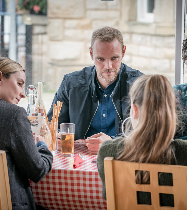 "The Bay - Vermutung": Lisa Armstrong (Morven Christie), Andy Warren (Joe Absolom), Rob Armstrong (Art Parkinson) und Abbie Armstrong (Imogen King) sitzen gemeinsam an einem Tisch in einem Restaurant. Lisa und Andy blicken zu Abbie, während Rob schmollend nach unten schaut.