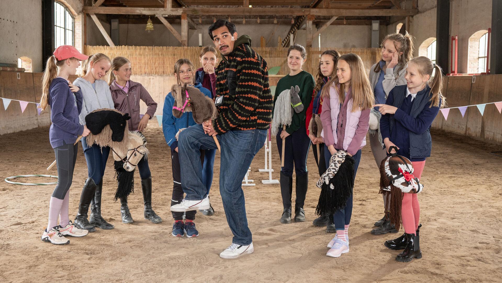 „SOKO Potsdam – Pferdemädchen“: In einer hellen Reithalle steht Samir Amari (Skandar Amini) zwischen einer Gruppe Mädchen, die Steckenpferde in den Händen halten. Umgeben von bunter Dekoration und Holzstangen versucht er, eine Reitbewegung nachzuahmen, während die Kinder amüsiert zusehen.