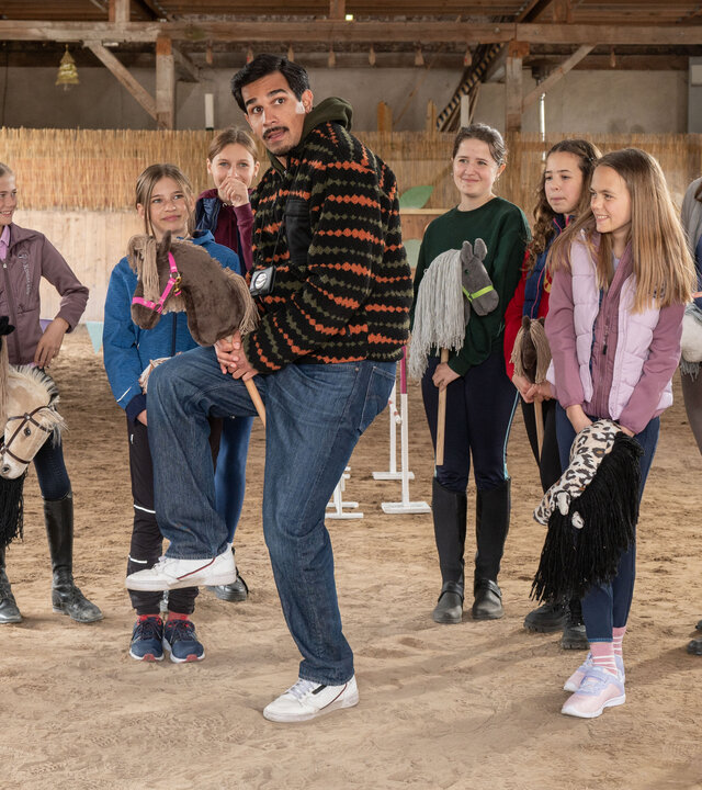 „SOKO Potsdam – Pferdemädchen“: In einer hellen Reithalle steht Samir Amari (Skandar Amini) zwischen einer Gruppe Mädchen, die Steckenpferde in den Händen halten. Umgeben von bunter Dekoration und Holzstangen versucht er, eine Reitbewegung nachzuahmen, während die Kinder amüsiert zusehen.