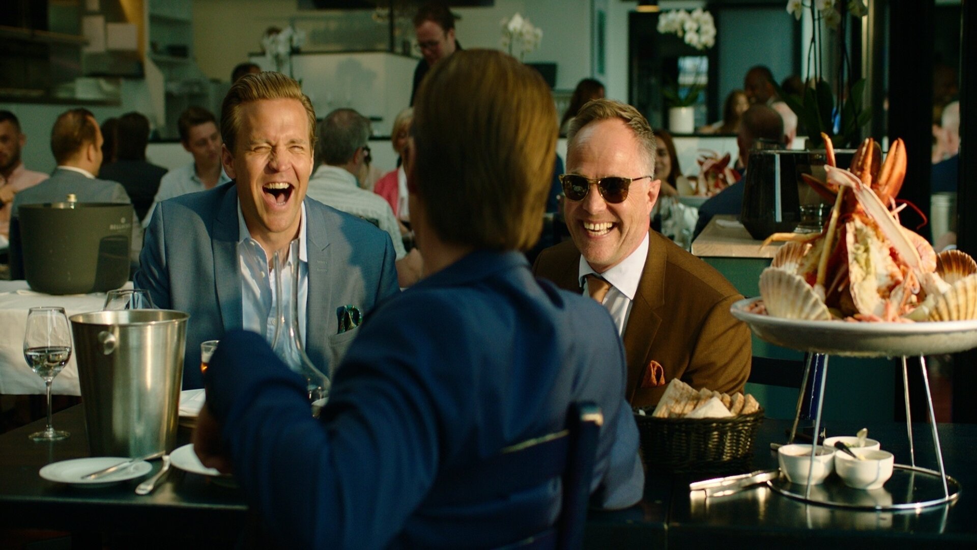 "Exit - Lazarus": Die Freunde Henrik (Tobias Santelmann), Jeppe (Jon Øigarden) und Adam (Simon J. Berger) sitzen lachend an einem Tisch in einem Restaurant.