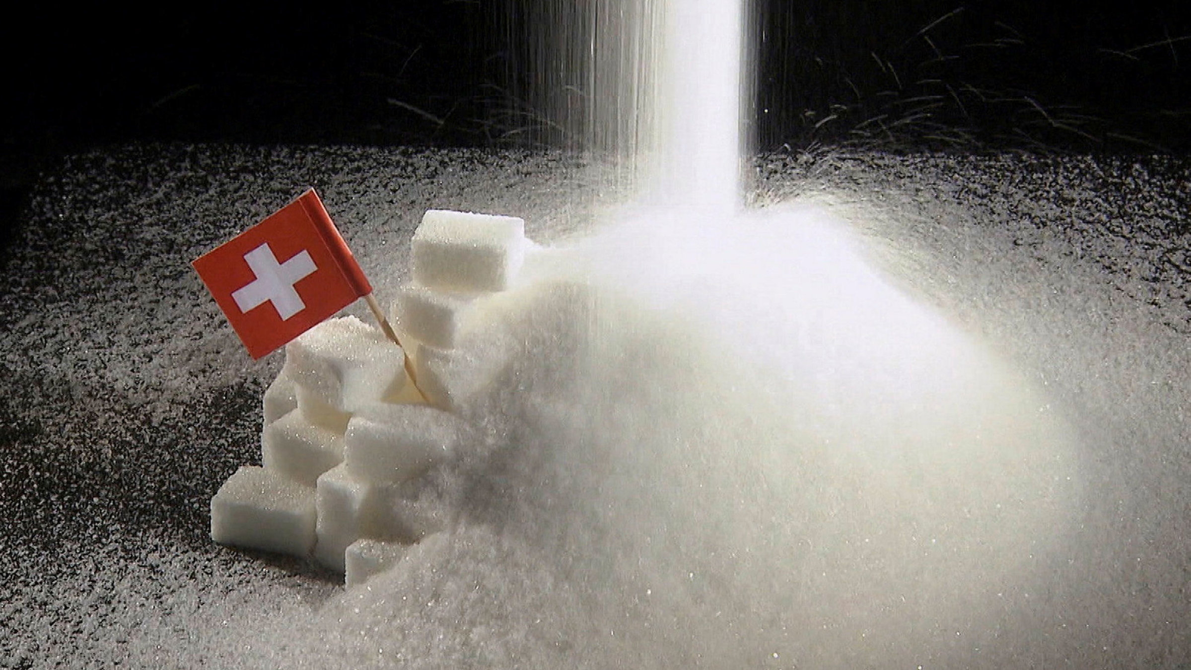 "Zucker – Die süße Droge": Zucker rieselt auf einen Haufen, in dem eine kleine schweizer Fahne steckt.