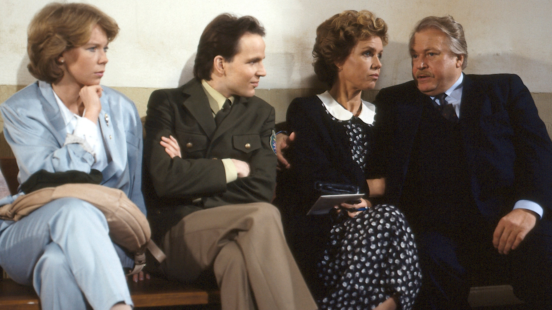 "Diese Drombusch - Der Prozeß": Tina Drombusch (Marion Kracht), Chris Drombusch (Mick Werup), Vera Drombusch (Witta Pohl) und Onkel Ludwig (Günter Strack) sitzen gemeinsam auf einer Bank vor einem Gerichtssaal.